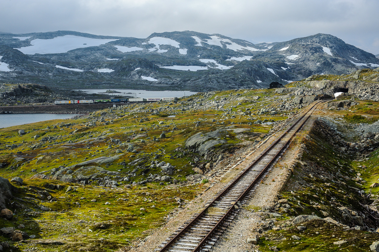 The line between Lågheller and Finse was replaced by a tunnel cutting through the mountain. The old line, climbing to 1349 meters, was difficult to keep open in the long winter. Here, freight train 5504 crosses Låghellervatnet before entering the tunnel. The old, dissused, line has branched of and climbs the hill in the foreground. 
